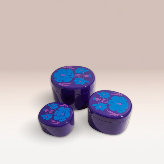 Lot de trois boites violettes foncées qui s'empilent comme des poupées russes. Des motifs floraux de couleur bleue et rose décorent le couvercle des trois boites.